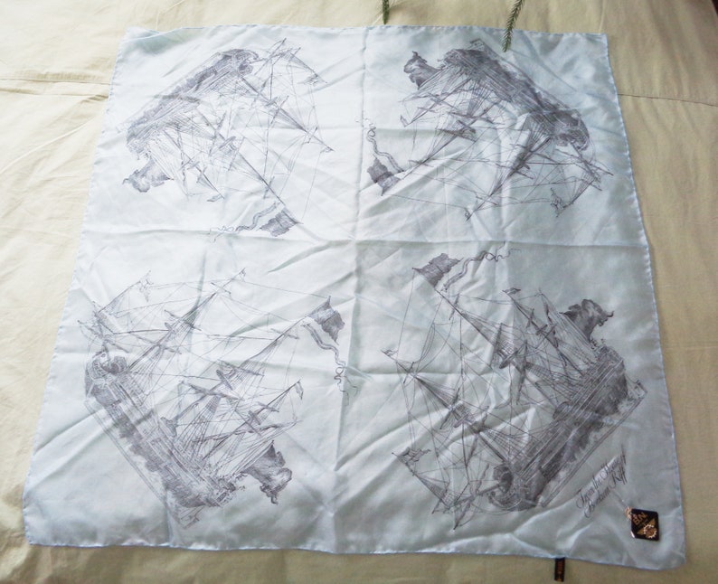 May include: A light blue square scarf featuring a repeating pattern of detailed gray ship illustrations. The scarf has a subtle sheen and a delicate appearance, with a small tag in the corner.