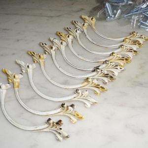May include: A collection of white, curved cabinet handles with ornate, gold-colored leaf accents. The handles are arranged in a row, with screws visible in the background. These decorative handles are ideal for furniture restoration or DIY projects.