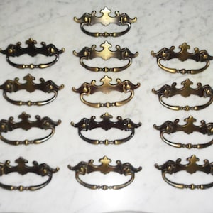May include: A set of 12 antique brass drawer pulls with ornate designs. Each pull has a curved shape with a decorative center and two side scrolls.