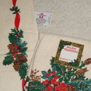 May include: Off-white holiday towels with a festive design. The towels feature a pattern of pinecones, holly, and red berries, accented with a red ribbon. The tag reads "Holiday Traditions".