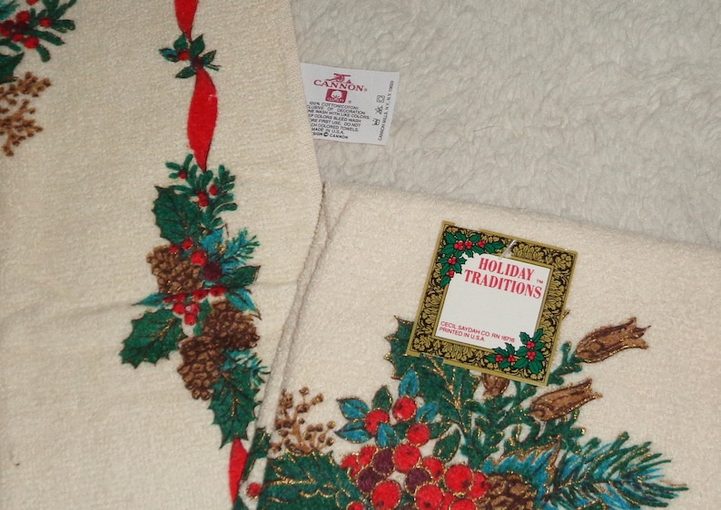 May include: Off-white holiday towels with a festive design. The towels feature a pattern of pinecones, holly, and red berries, accented with a red ribbon. The tag reads "Holiday Traditions".