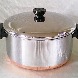 以下が含まれることがあります： 蓋と黒いハンドルが付いたステンレス製の調理鍋。鍋は銅色のベースと蓋に黒いノブが付いています。 キッチン用品。