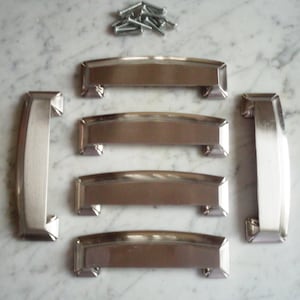 Puede incluir: Cinco tiradores de armario plateados con forma rectangular ligeramente curvada y acabado cepillado. Los tiradores están dispuestos en fila, con el tirador superior ligeramente desplazado hacia la derecha.