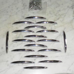 Puede incluir: Un juego de 20 tiradores de armario cromados con un diseño curvo en forma de barco. Los tiradores están dispuestos en filas sobre una superficie blanca.