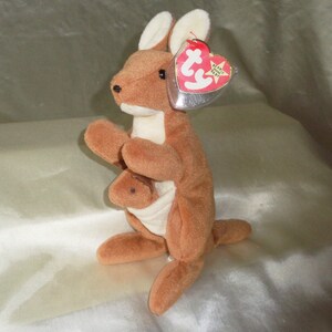 May include: A brown and white plush kangaroo with a joey in its pouch. The kangaroo has a red Ty Beanie Baby tag attached to its ear. The tag says "Ty Beanie Babies".