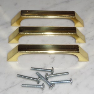Puede incluir: Tres tiradores de armario de color dorado con forma rectangular y bordes redondeados. Los tiradores están apilados, con el tirador superior ligeramente desplazado hacia la derecha. Los tiradores se acompañan de un juego de seis tornillos plateados.