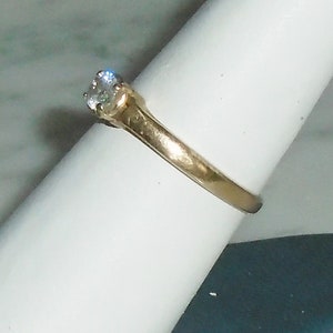 May include: A gold-colored ring with a single, clear, round-cut gemstone. The ring is set on a white surface, showcasing the band's design and the brilliance of the stone. The ring is a classic style, suitable for various occasions.