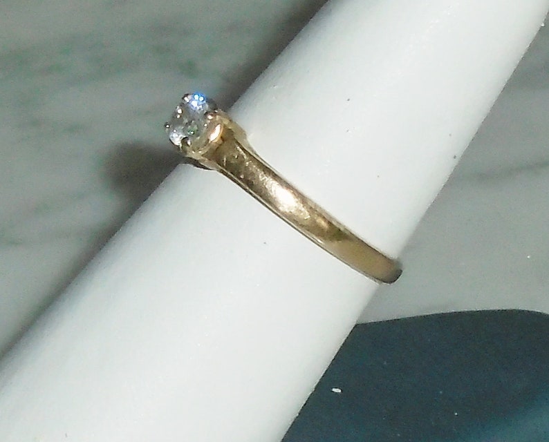 May include: A gold-colored ring with a single, clear, round-cut gemstone. The ring is set on a white surface, showcasing the band's design and the brilliance of the stone. The ring is a classic style, suitable for various occasions.