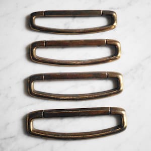 Puede incluir: Cuatro lazos de cinturón o soportes de correa de metal dorado. Los lazos son rectangulares con bordes redondeados y una ligera curva en el centro.