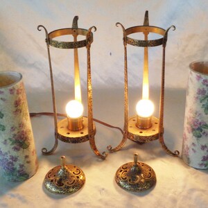 May include: Two antique brass table lamps with floral patterned shades. The lamps have a simple design with a round base and a tall, cylindrical shade. The shades are made of a white material with a floral pattern in shades of purple and green.