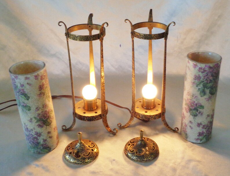 May include: Two antique brass table lamps with floral patterned shades. The lamps have a simple design with a round base and a tall, cylindrical shade. The shades are made of a white material with a floral pattern in shades of purple and green.