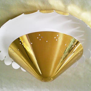 Może przedstawiać: Zabytkowa lampa sufitowa w stylu mid-century modern. Lampa ma złoty, stożkowaty środek z małymi perforacjami i białą, karbowaną krawędzią. Przeznaczona do montażu na suficie.