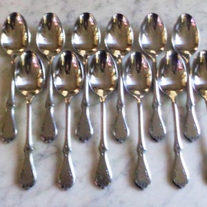 Puede incluir: Un conjunto de doce cucharas de plata, cada una con un cuenco redondeado y un mango adornado. Las cucharas están dispuestas en filas, reflejando la luz. Las cucharas son probablemente para servir o comer.