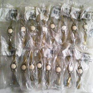 May include: A collection of sixteen individual Amerock cabinet pulls, each encased in clear plastic packaging. The pulls feature an antique brass finish with a central circular cream-colored inlay, decorative scrollwork, and include mounting screws.
