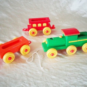 Puede incluir: Un tren de juguete de madera con una locomotora verde, un vagón de cola rojo y un vagón rojo. El tren tiene ruedas amarillas y un techo rojo. El tren está sobre una superficie blanca.