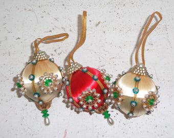 May include: Three vintage Christmas ornaments. Two are cream-colored, and one is red. Each ornament is decorated with pearls, green and blue accents, and a gold-colored hanging loop. The ornaments have a classic, festive design.