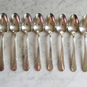 May include: Seven silver spoons arranged in a row. Each spoon has a rounded bowl and a long, slender handle. The spoons reflect light, creating a bright, shiny appearance. The spoons are likely for serving or eating.