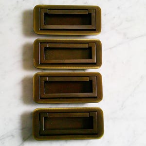 Puede incluir: Cuatro tiradores de cajón empotrados rectangulares de color bronce. Cada tirador tiene un marco rectangular con un asa empotrada. Los tiradores están dispuestos verticalmente sobre un fondo de mármol blanco. Herrajes para muebles.