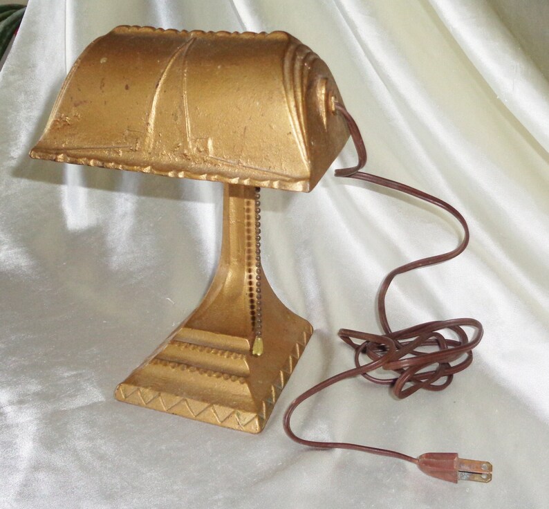 May include: A gold-colored metal desk lamp with a curved shade and a chain to adjust the height. The lamp has a brown cord and plug.