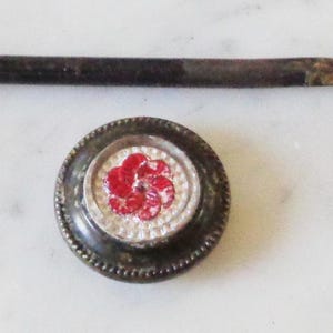 May include: A vintage metal button with a red and white flower design. The button is attached to a long, thin metal rod.