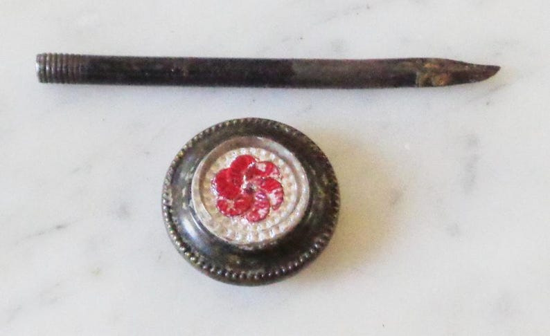 May include: A vintage metal button with a red and white flower design. The button is attached to a long, thin metal rod.