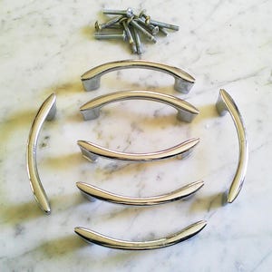 Könnte beinhalten: Eine Sammlung von sechs polierten Chrom-Schrankgriffen und einem kleinen Haufen Schrauben. Die Griffe sind gebogen und haben eine glatte, reflektierende Oberfläche. Die Hardware ist auf einer weiß-grauen Marmoroberfläche angeordnet.