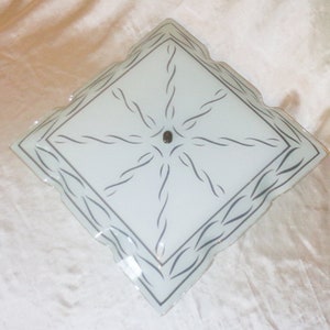 May include: A white square ceiling light fixture with a decorative, wavy, black design. The light fixture has a silver center.