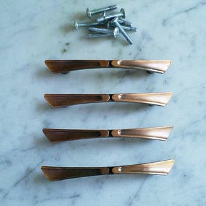 Puede incluir: Cuatro tiradores de armario de color cobre con diseño de lazo, junto con un montón de tornillos plateados. Los tiradores están dispuestos en fila, mostrando su acabado metálico y forma decorativa. Ideal para renovaciones de muebles.