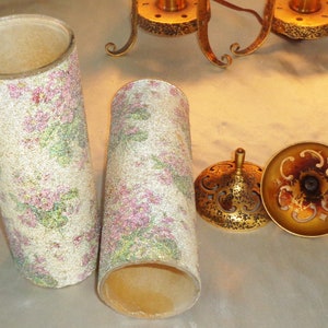 May include: Two white lampshades with a pink and green floral pattern. The lampshades are made of a textured material and have a gold metal base.