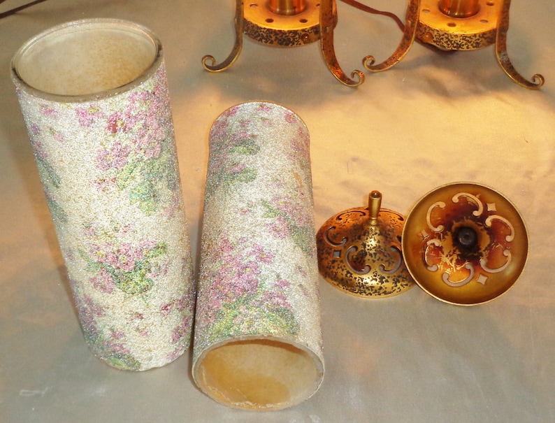 May include: Two white lampshades with a pink and green floral pattern. The lampshades are made of a textured material and have a gold metal base.