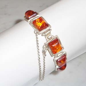 May include: A silver bracelet featuring rectangular amber stones. The amber stones are a warm orange-brown color and are set in a silver frame. A delicate silver chain is attached to the bracelet for added security and style. The bracelet is a piece of jewelry.