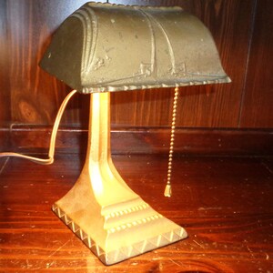 May include: A gold-toned metal desk lamp with a decorative, scalloped shade and a pull chain. The lamp has a simple, elegant design.