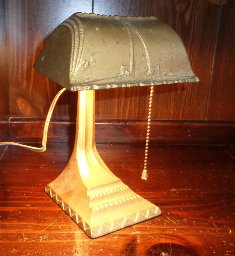 May include: A gold-toned metal desk lamp with a decorative, scalloped shade and a pull chain. The lamp has a simple, elegant design.