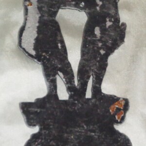 Puede incluir: Una silueta de metal plateado de dos figuras de pie una al lado de la otra sobre una base de metal negro. Las figuras se toman de la mano y miran hacia adelante. La base tiene un dise&ntilde;o decorativo.
