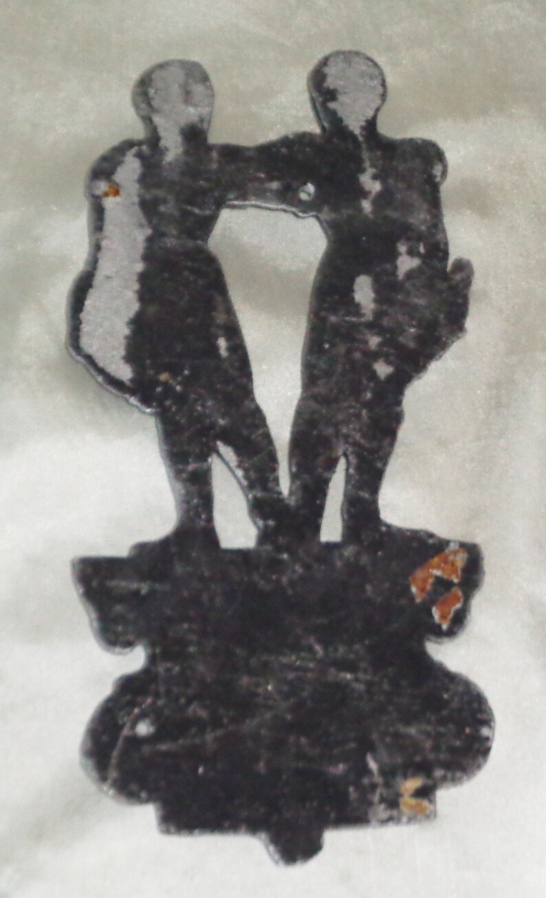 Puede incluir: Una silueta de metal plateado de dos figuras de pie una al lado de la otra sobre una base de metal negro. Las figuras se toman de la mano y miran hacia adelante. La base tiene un dise&ntilde;o decorativo.