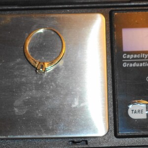 May include: A gold ring with a small diamond is placed on a digital scale. The scale displays a weight of 2.2 grams. The scale has a capacity of 600g and a graduation of 0.1g.