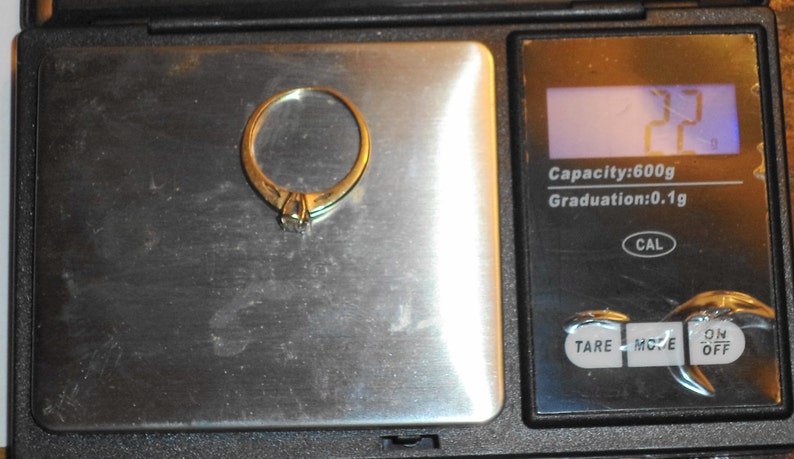 May include: A gold ring with a small diamond is placed on a digital scale. The scale displays a weight of 2.2 grams. The scale has a capacity of 600g and a graduation of 0.1g.