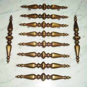 May include: Ten antique brass cabinet knobs with a decorative, ornate design. The knobs have a spindle shape with a rounded top and a pointed bottom.