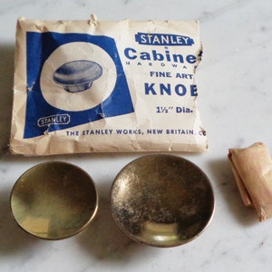 May include: A packet of Stanley Cabinet Hardware Fine Art Knobs, 1 1/2 inches in diameter. The packet is brown with blue text and a picture of a brass knob. Two brass knobs are shown outside of the packet.