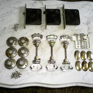 Puede incluir: Un surtido de herrajes antiguos para puertas, que incluye tres pomos de puerta de cristal con vástagos metálicos, varias rosetas de latón y tres pestillos de puerta de metal negro. También se incluyen varios tornillos pequeños y placas metálicas.