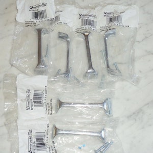Könnte beinhalten: Sechs silberne Schrankgriffe mit Schrauben in einzelnen Plastikverpackungen. Die Verpackung trägt den Markennamen Amerock und die Produktnummer BP76225-26.