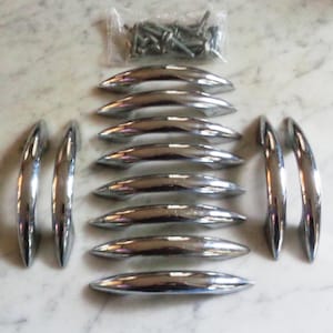 Puede incluir: Un conjunto de once tiradores de armario cromados pulidos, dispuestos sobre una superficie clara. Los tiradores son curvos con un diseño aerodinámico. También se ve una pequeña bolsa de tornillos.