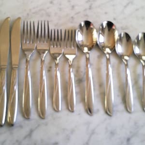 May include: A set of silver-colored flatware, including knives, forks, and spoons, arranged on a marble surface. The knives have a curved blade, and the forks have four tines. The spoons are oval-shaped. The flatware has a simple, elegant design.
