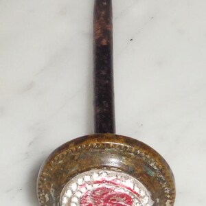 May include: A vintage brass tack with a red and white enamel flower design. The tack has a long, thin, rusty metal shaft.