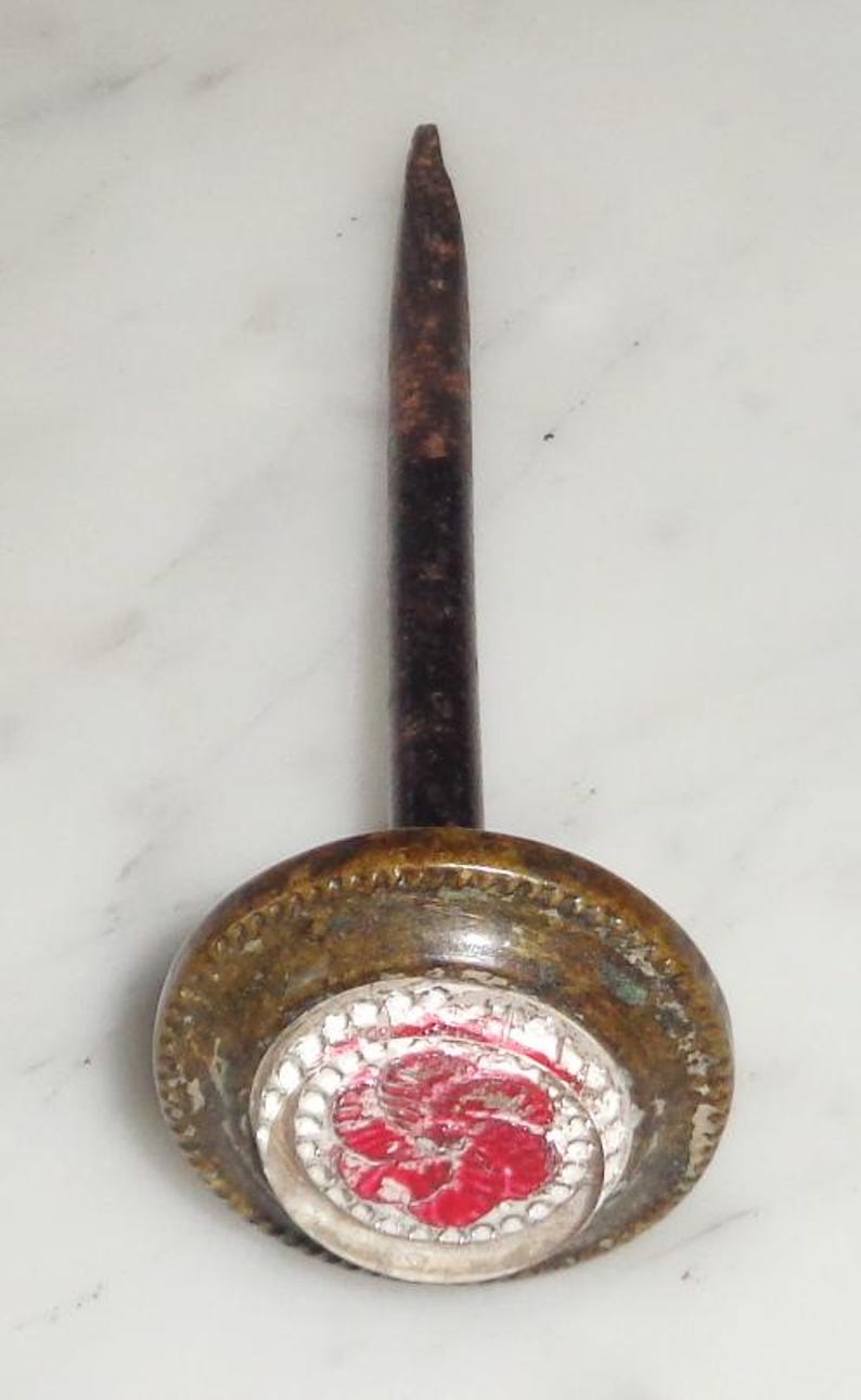 May include: A vintage brass tack with a red and white enamel flower design. The tack has a long, thin, rusty metal shaft.