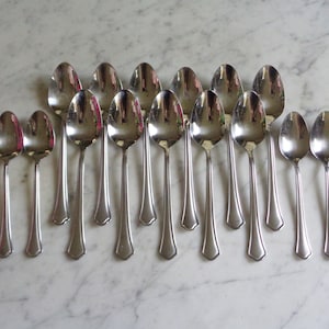May include: A set of 15 silver spoons with a rounded bowl and a slightly flared handle. The spoons are arranged in a row on a white marble surface.