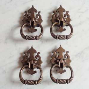 Puede incluir: Cuatro tiradores de puerta de armario de latón antiguo con diseños ornamentados. Cada tirador tiene una manija de anillo y una parte superior decorativa.