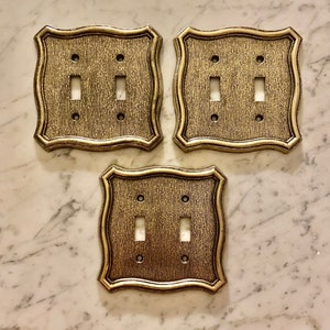 Puede incluir: Tres tapas de interruptor de luz de color dorado con un diseño decorativo y ornamentado. Cada tapa tiene dos interruptores de palanca.