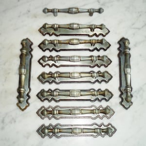 Puede incluir: Conjunto de diez tiradores de cajones antiguos de color plateado con diseños ornamentados. Los tiradores tienen un acabado ligeramente envejecido y están dispuestos en dos filas sobre una superficie de mármol blanco.