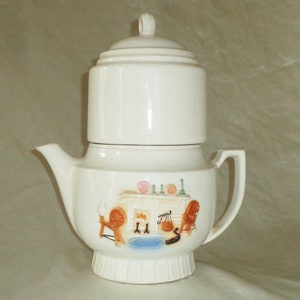 Puede incluir: Una tetera vintage de cerámica blanca con tapa a juego. La tetera presenta una escena decorativa de una cocina con una rueca, una balanza y una silla. La tetera tiene un asa y un pico.